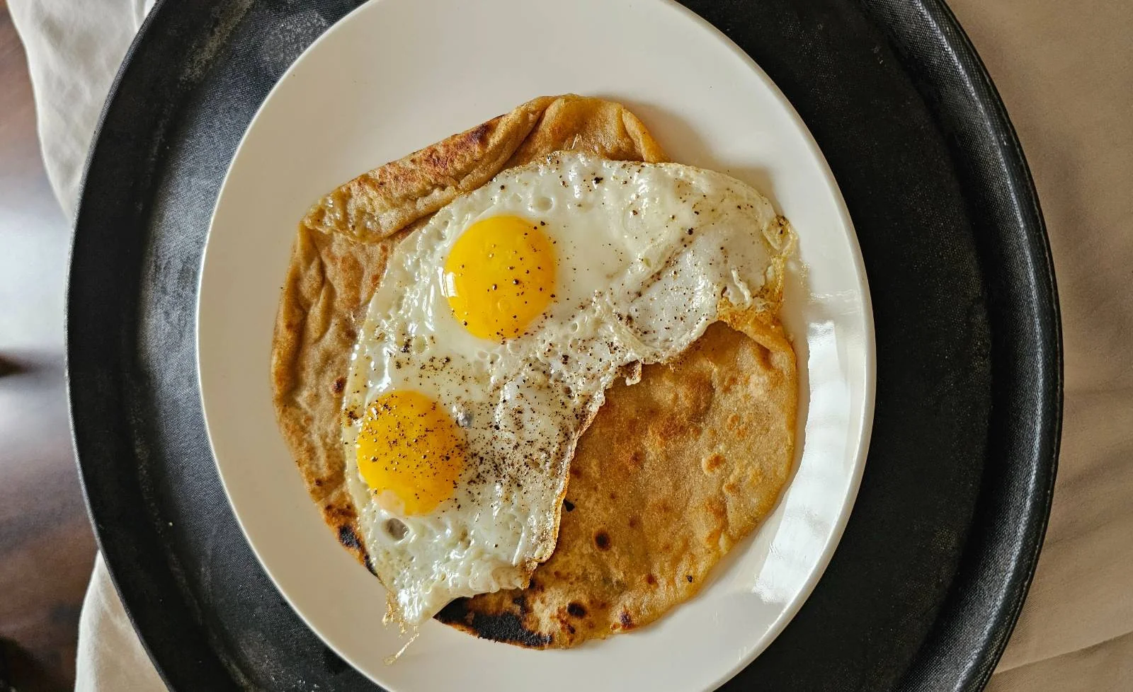 A white plate with two sunny-side-up eggs placed on top of a flatbread, served on a dark tray.