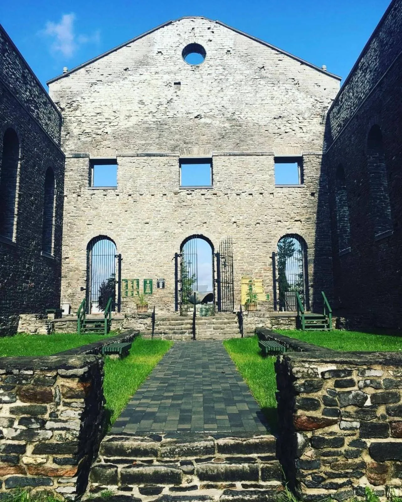 Stone ruins of a large building with a triangular roof, three tall arched windows, and a pathway with steps leading to the entrance, surrounded by grass and blue sky above.