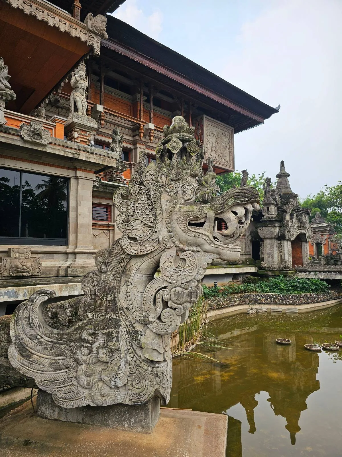 A large stone dragon sculpture stands beside a shallow pond with a traditional Balinese building in the background, featuring ornate carvings and a tiered roof. Lush greenery is visible, enhancing the tranquil atmosphere.