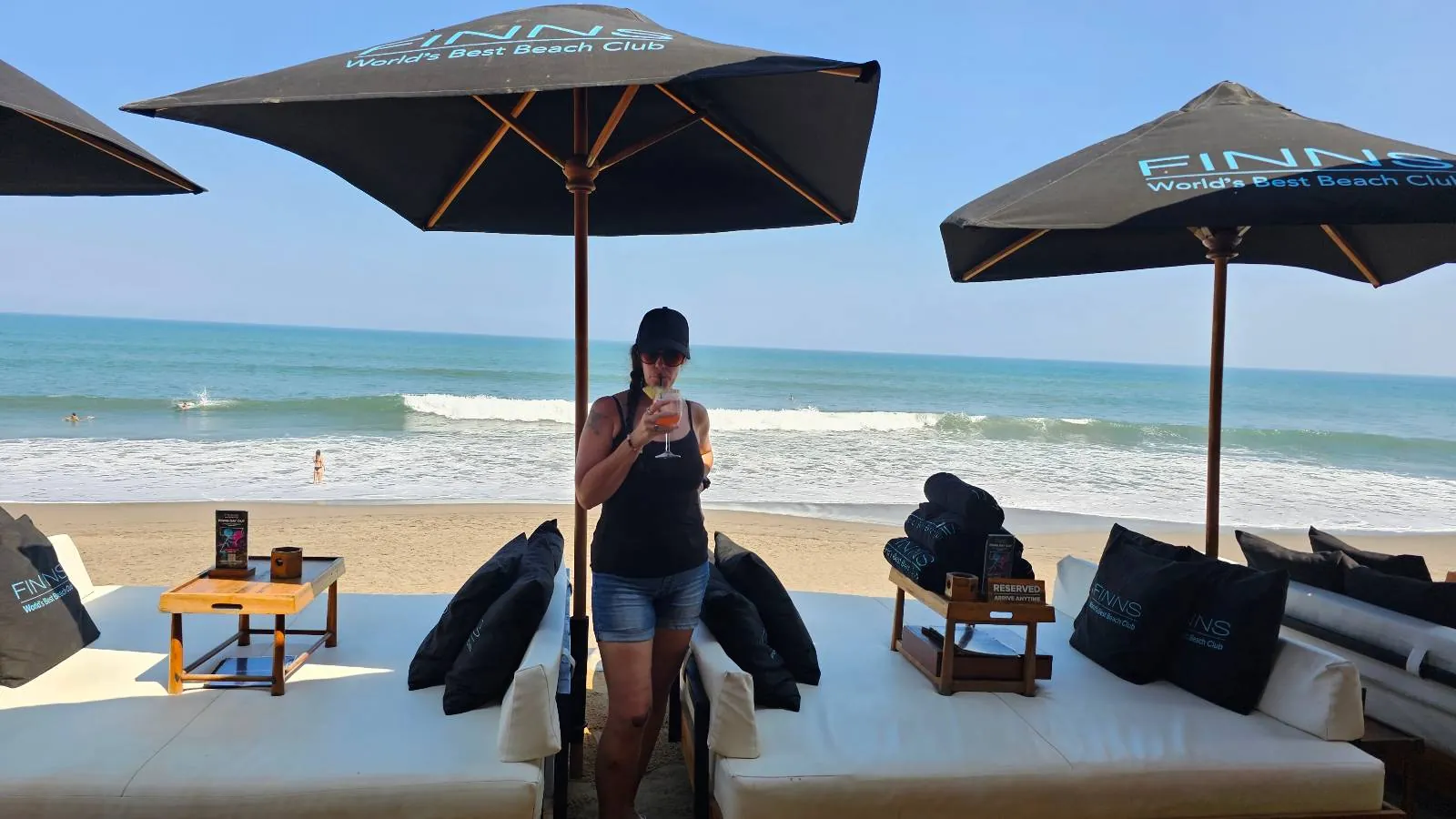 A person in a black hat and tank top stands between two lounge chairs under large umbrellas at a beach. The ocean waves are visible in the background under a clear blue sky.