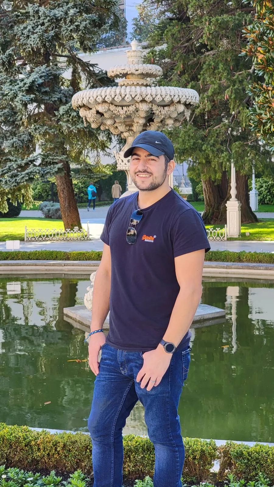 A man wearing a black cap, black t-shirt, and jeans stands in front of a decorative fountain in a garden with green trees and manicured hedges in the background.