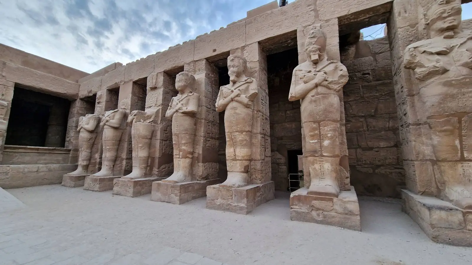 A row of ancient stone statues stands tall against a backdrop of weathered Egyptian temple walls. The statues have crossed arms and wear traditional headdresses, positioned in a semi-enclosed open-air area under a partly cloudy sky.