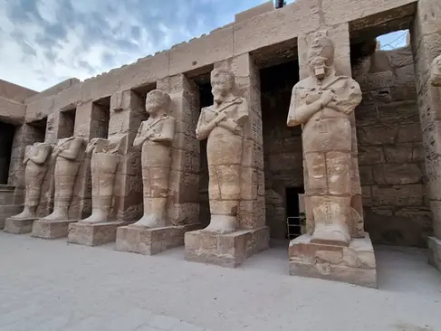 A row of ancient stone statues stands tall against a backdrop of weathered Egyptian temple walls. The statues have crossed arms and wear traditional headdresses, positioned in a semi-enclosed open-air area under a partly cloudy sky.
