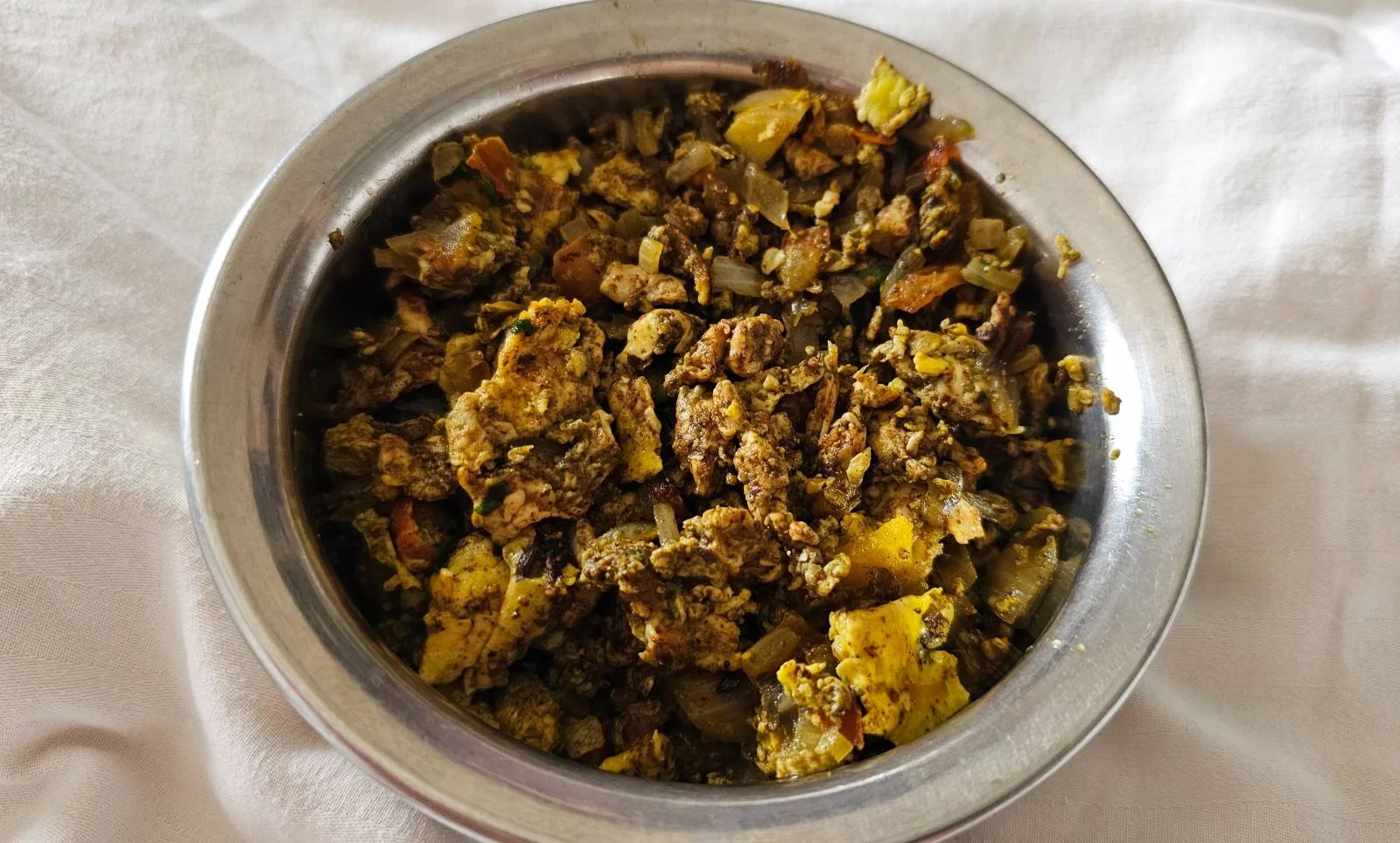 A round metal bowl filled with a scrambled dish that appears to contain diced vegetables and eggs, placed on a white cloth background.