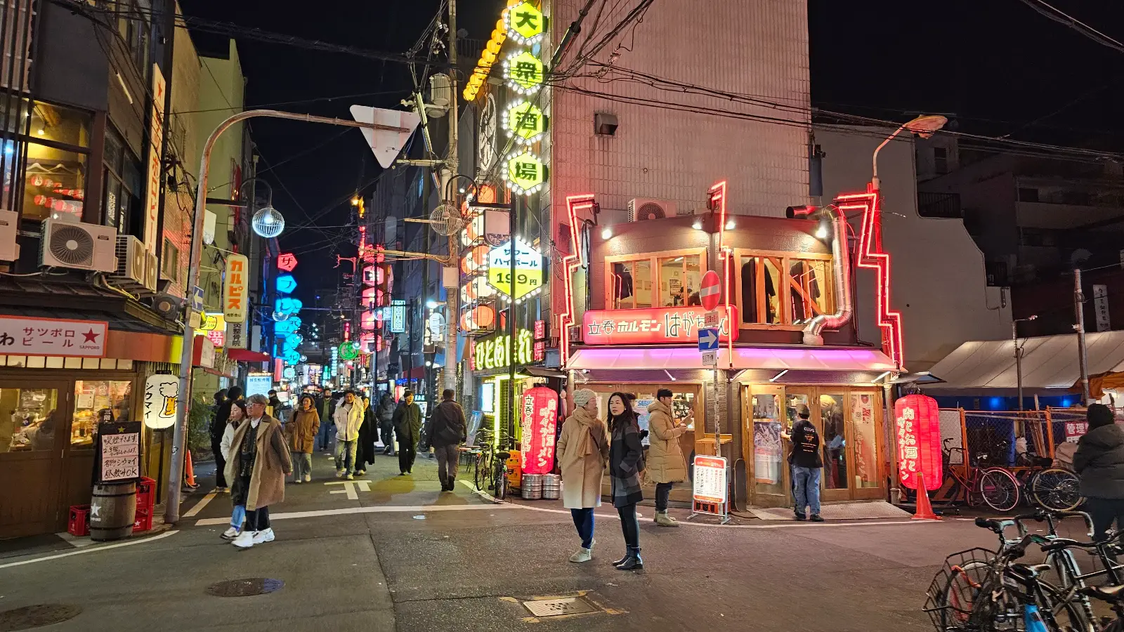 A lively city street at night with people walking, colorful neon signs, and lights illuminating shops and restaurants in an urban area.