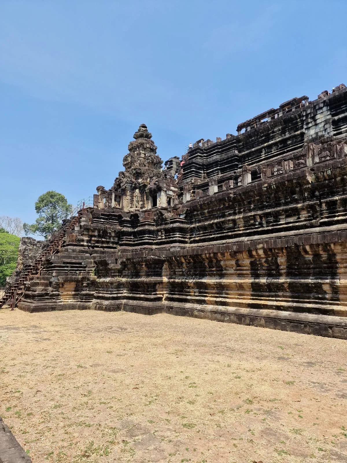 Ancient stone temple with multiple tiers and detailed carvings, set against a clear blue sky and surrounded by dry grass and sparse trees.