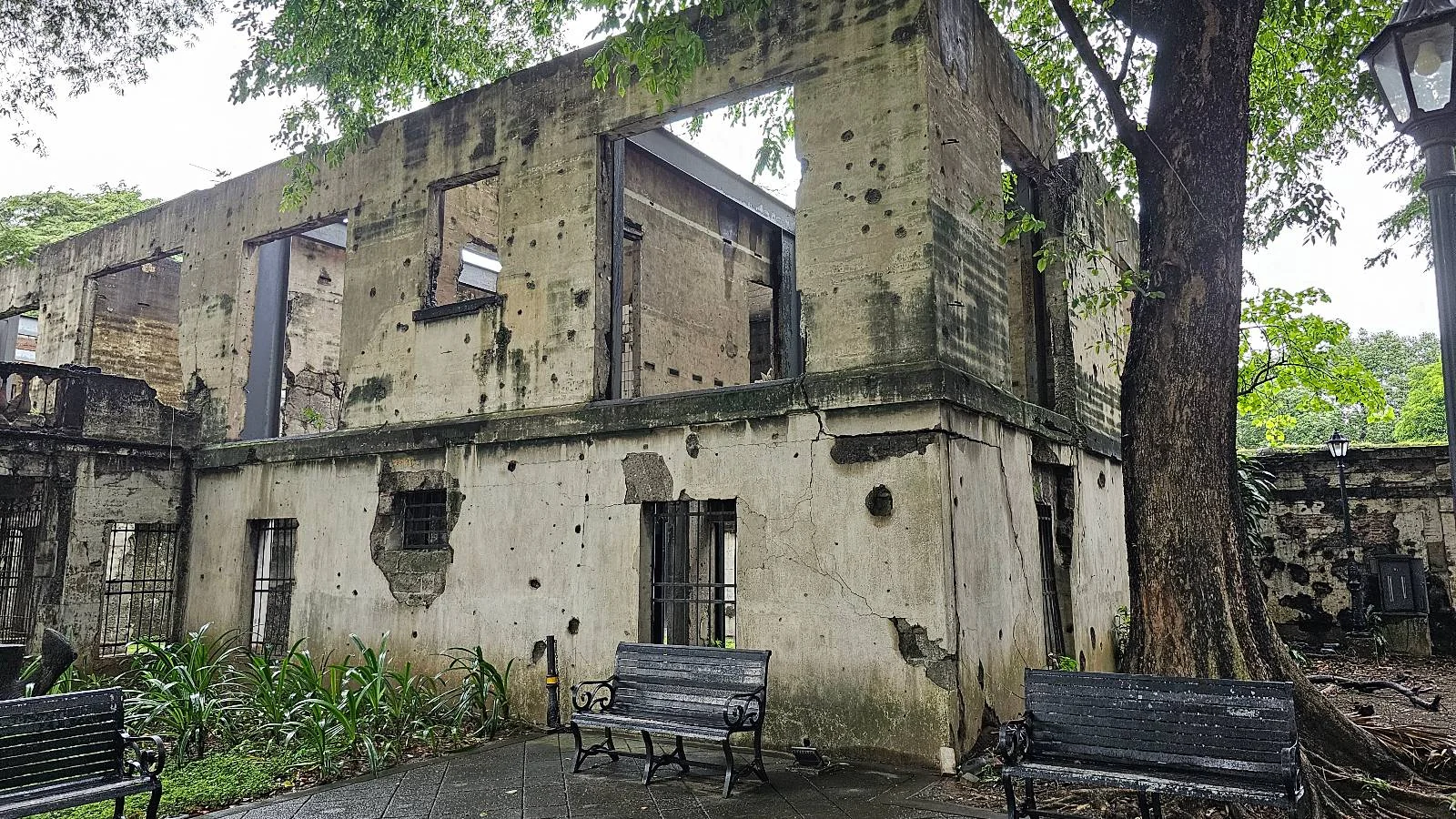 A weathered, bullet-riddled stone building with no roof stands under a tree. Empty window frames and benches are visible, with greenery around. The building looks historic and is set in a park-like environment.