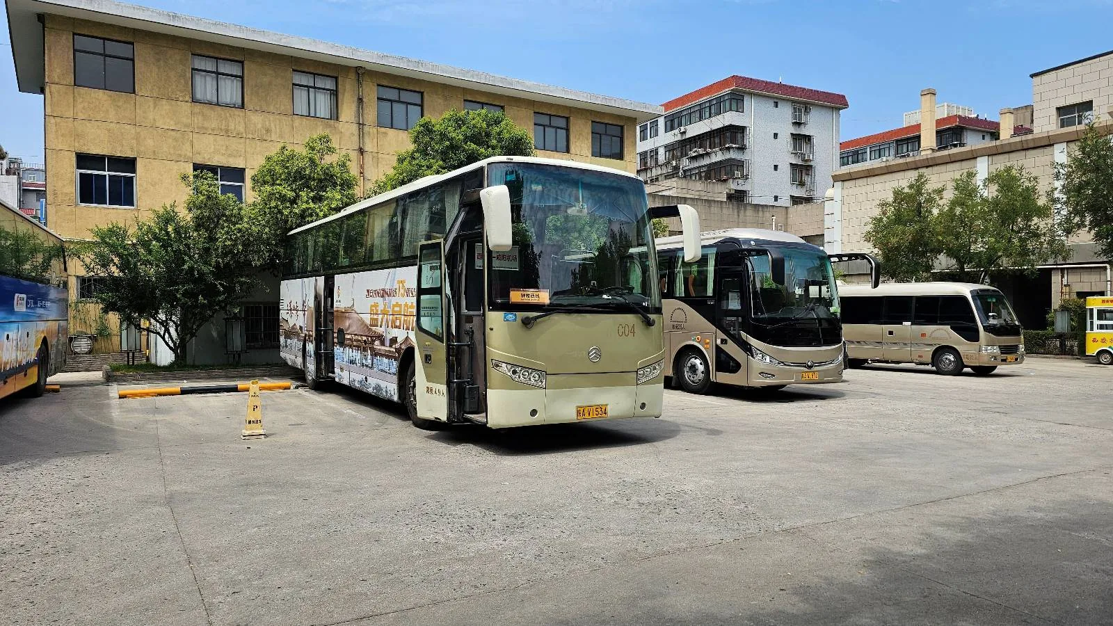 Several buses are parked in a paved lot near a yellow multi-story building with large windows, surrounded by trees and other buildings under a clear blue sky.