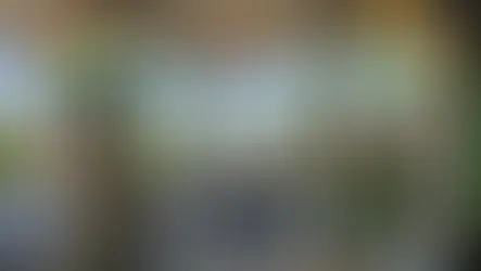 Rooftop terrace with tables and chairs overlooking Udaipur cityscape. Decorative columns, plants, and "Dreamyard Udaipur Hostel" sign. Bright daylight.