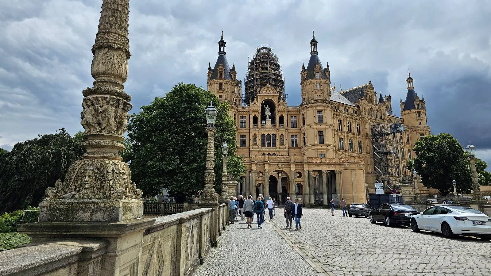 Historic castle with ornate towers and a large dome sits under a cloudy sky; people walk along a cobblestone path, and cars are parked on the side. Decorative stone pillars line the walkway.