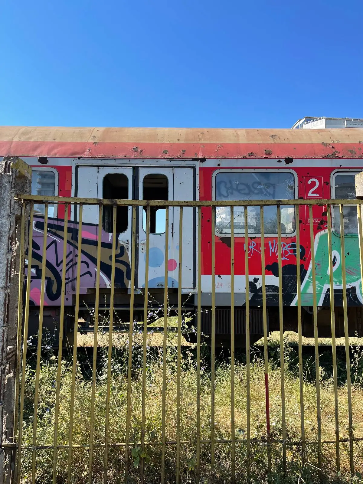 A red and white train car with graffiti on its side stands on overgrown tracks behind a metal fence under a clear blue sky.