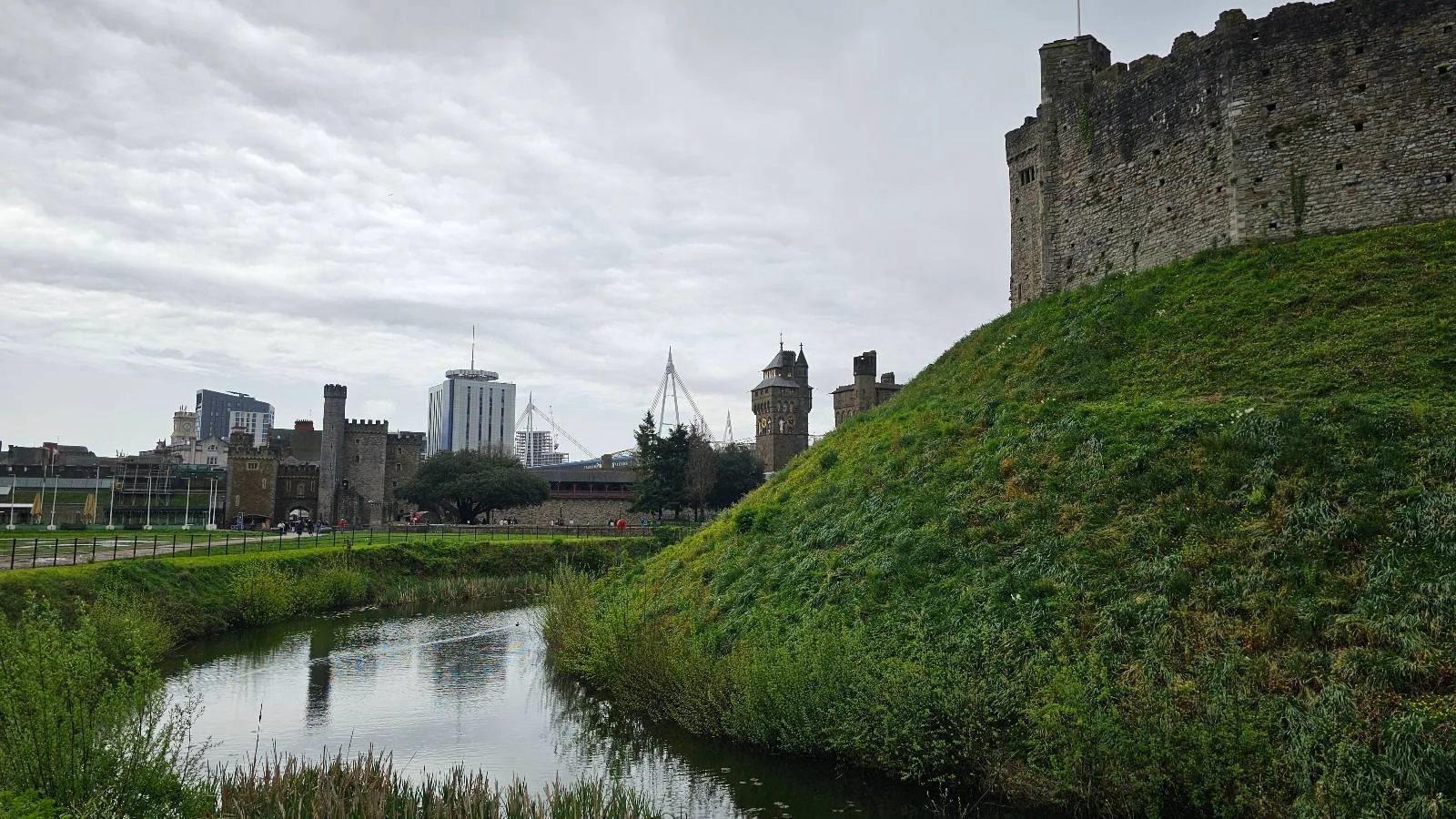 A scenic view of a medieval castle atop a lush green hill, with a moat in the foreground and modern skyscrapers in the distance under a cloudy sky.