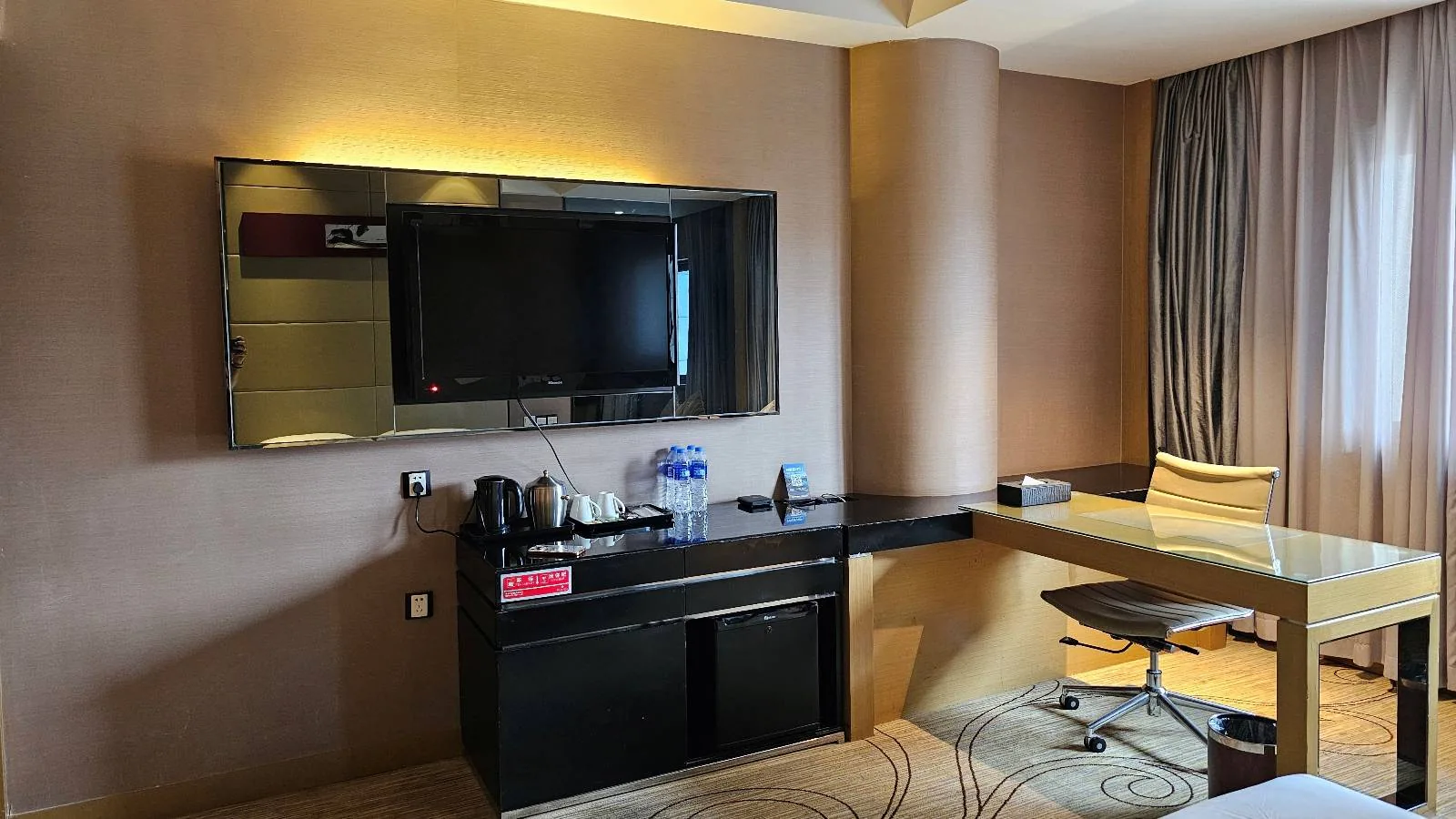 A modern hotel room features a wall-mounted flat-screen TV, a desk with a chair, a minibar with drinks, and large windows with curtains letting in natural light.