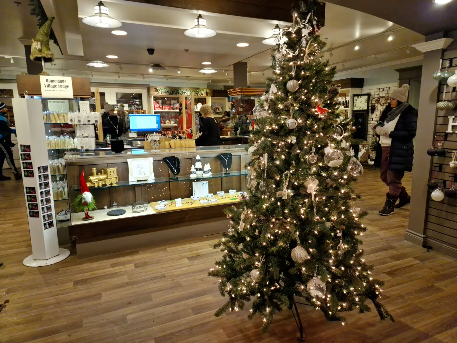 A decorated Christmas tree with white lights and ornaments stands in the center of a store with wooden floors and shelves of products in the background.