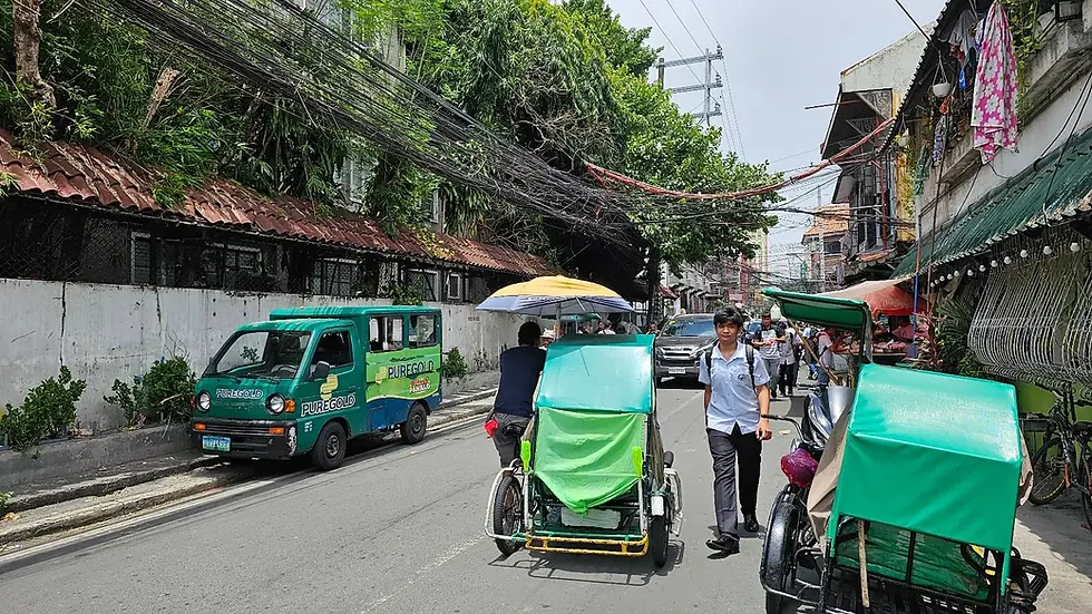 Pedestrians and drivers navigate a busy street lined with tangled utility wires overhead and various vehicles including a green public van and pedal-powered rickshaws.
