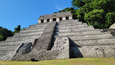 The image showcases an ancient Maya stepped pyramid nestled in a jungle setting under a clear blue sky. The grey stone structure, with its multiple terraces, ascends to a small building at the top. Dense green foliage envelops this splendid testament to South American history and archaeology.