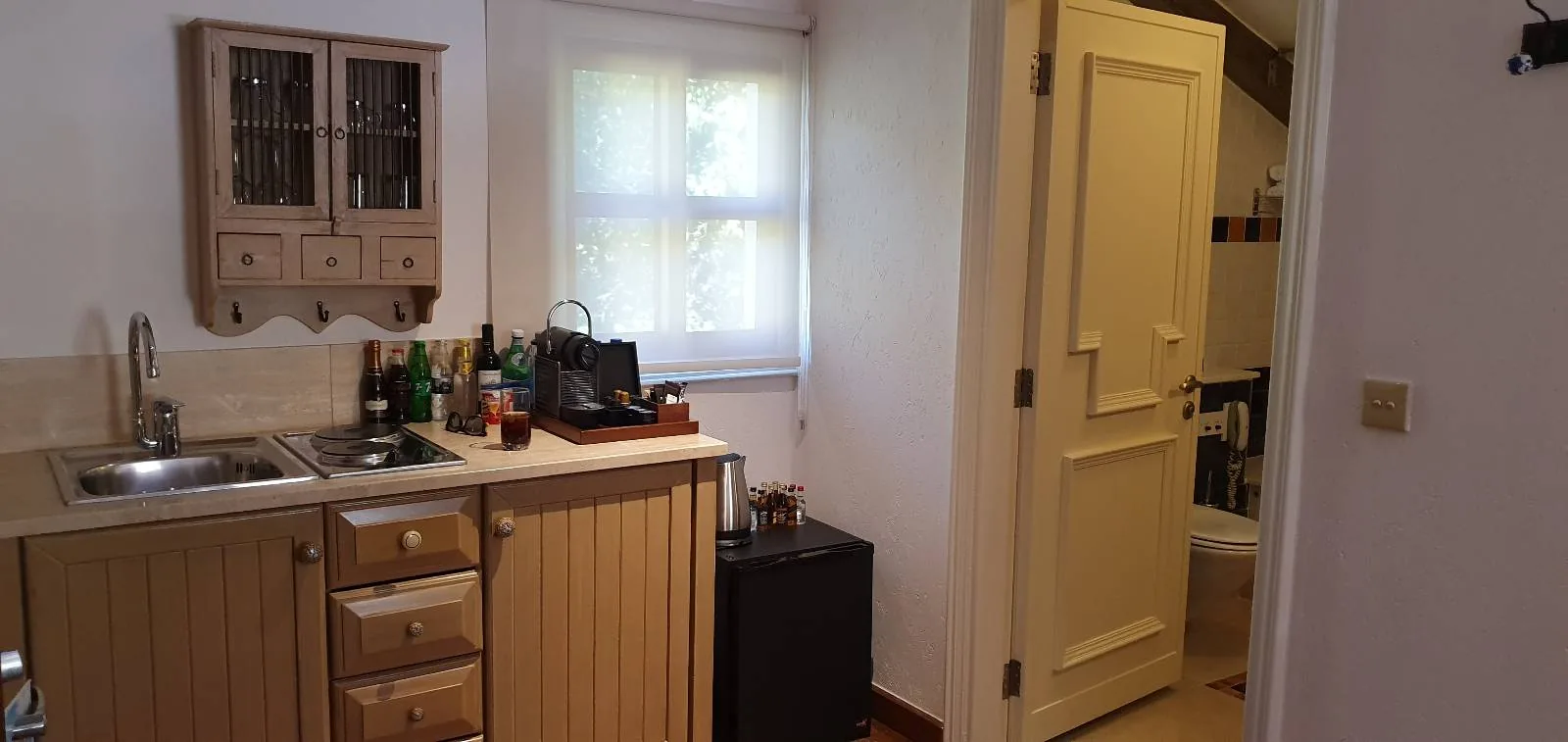 A small kitchen area with wooden cabinets, a sink, and various items on the counter. A window with sheer curtains is above the counter. To the right, there is an open door leading to a bathroom with visible fixtures.