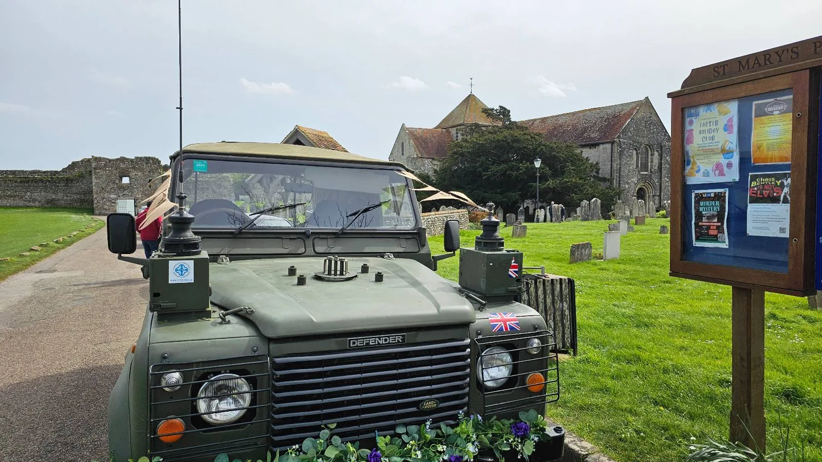 A green land rover defender parked beside a cemetery and an old church, with informational signage in the foreground.