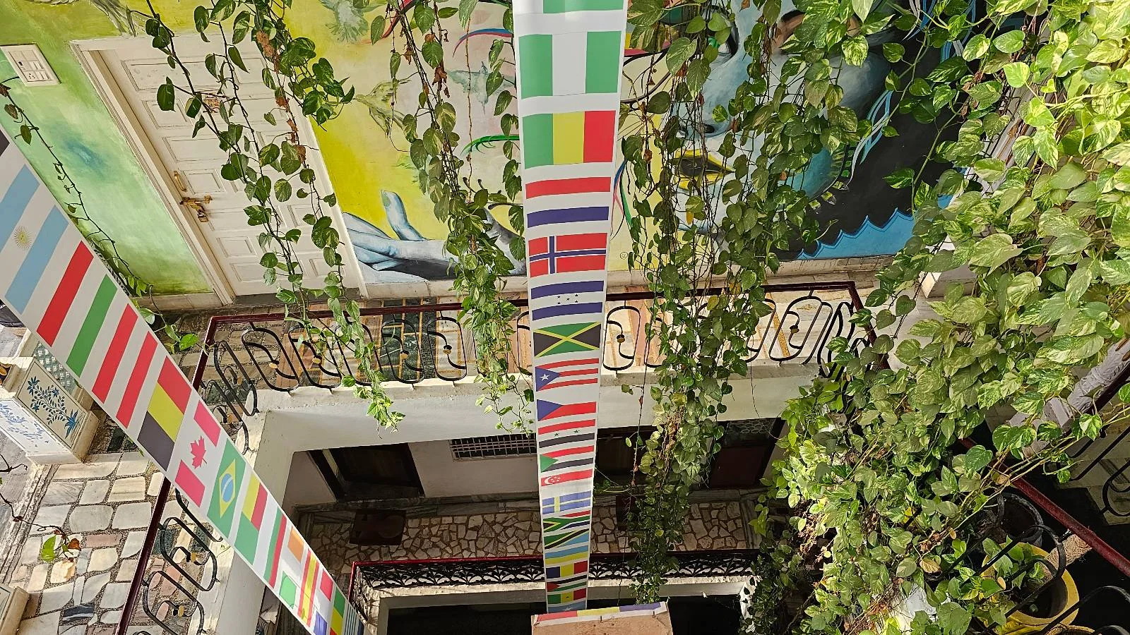 A view looking up inside a building decorated with hanging strands of international flags and lush green plants, with balconies and natural light visible.