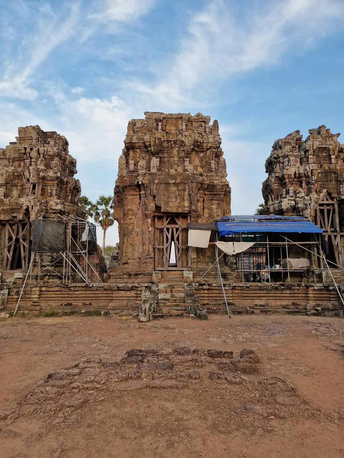 Ancient stone temple ruins with three towers, partially covered by a tarp and scaffolding, under a blue sky with wispy clouds. The ground is dry and reddish, and a palm tree is visible between the towers.