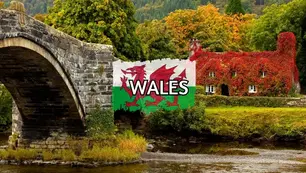 A picturesque stone bridge spans a river in an autumnal landscape, with a cottage covered in red ivy in the background. The image is overlaid with the Welsh flag and the word "Wales.