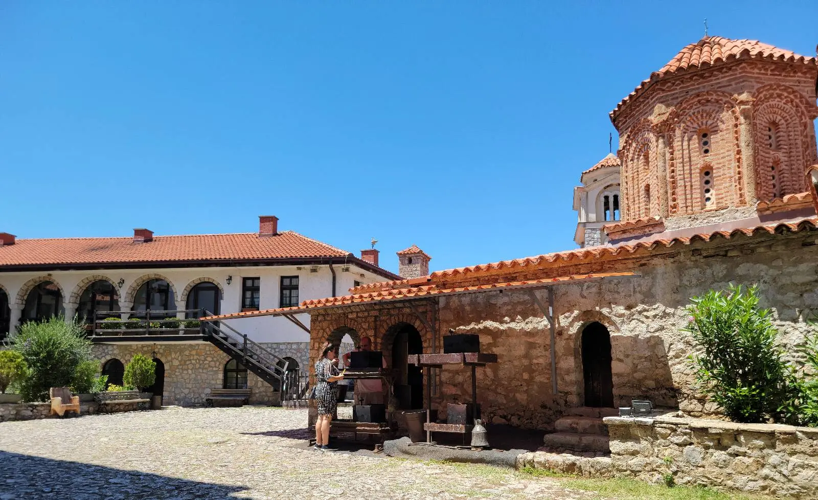 A historic courtyard featuring stone buildings under a clear blue sky. On the right, a red-roofed domed structure, while the left shows a building with arched windows and a staircase. Cobblestone ground and a few plants are visible.