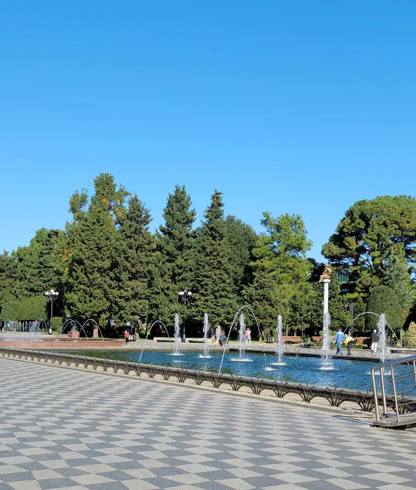 Chequered square paving leading up to a rectangular pool fountain with green trees in the background.