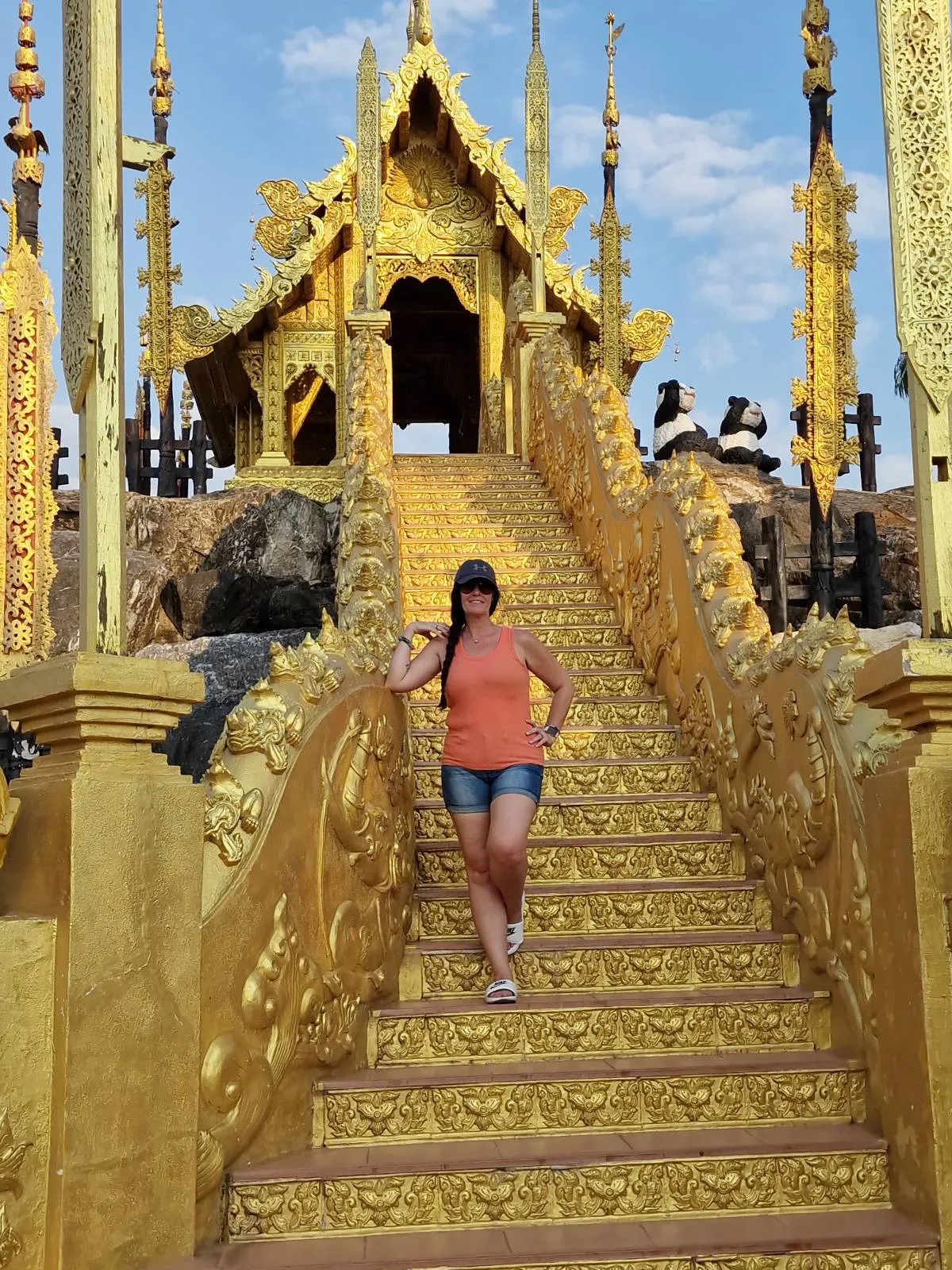 A person stands on ornate golden stairs leading to an intricately decorated temple with tall, spire-like structures. The sky is blue with some clouds, and the person is wearing casual clothes, posing for the photo.