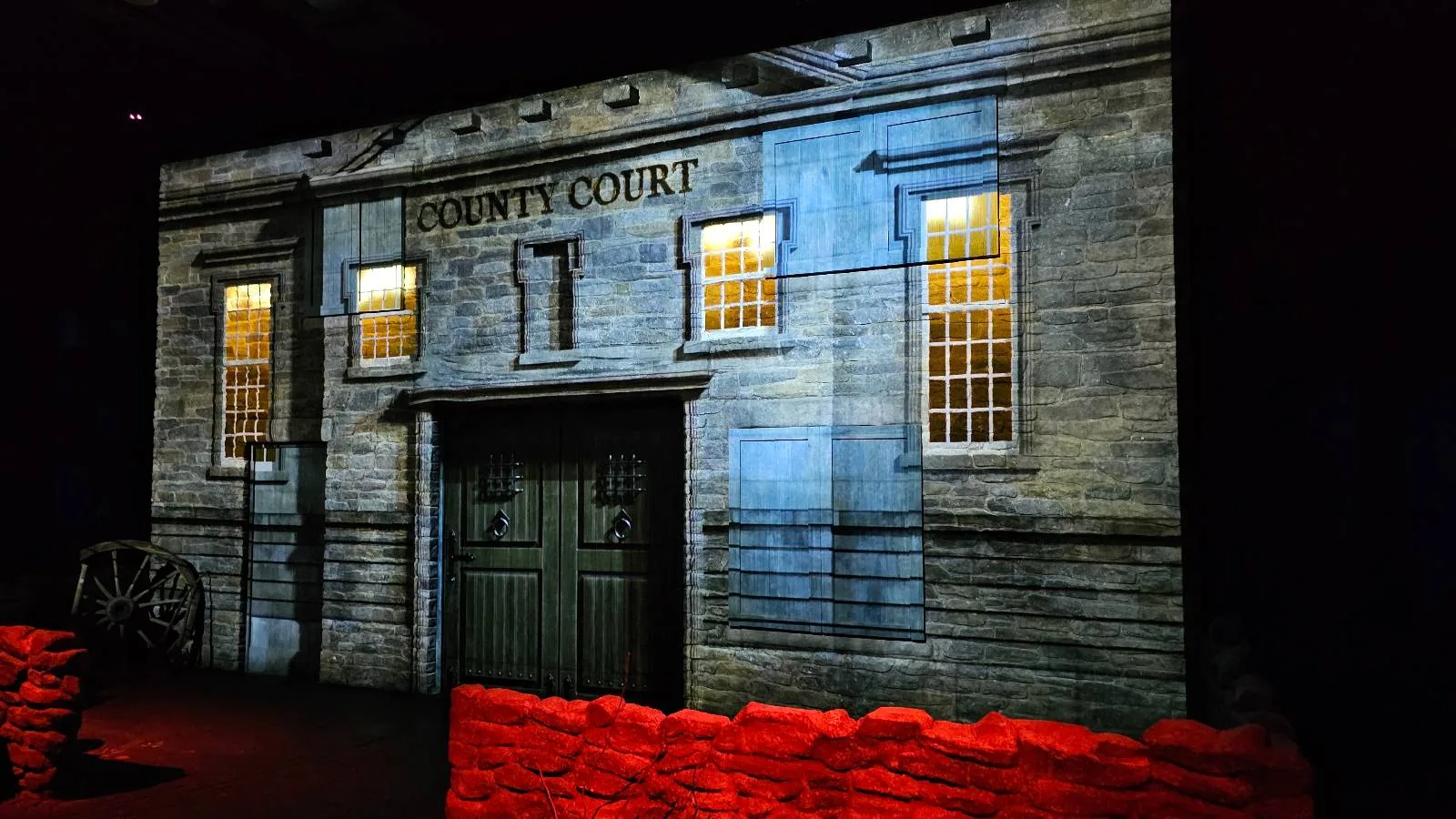 A dimly lit scene of an old county courthouse facade with glowing windows, double doors, and "County Court" written above. The foreground features a low red-lit barrier and a wagon wheel on the left.