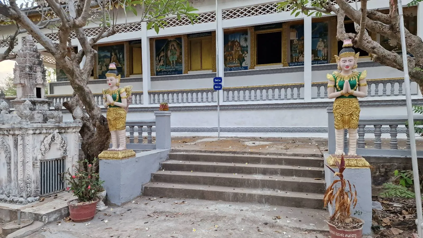 A sandy open area with a traditional building on the left, colorful flags, trees, a small shrine or structure in the center, and a white car parked on the right under a cloudy early evening sky.