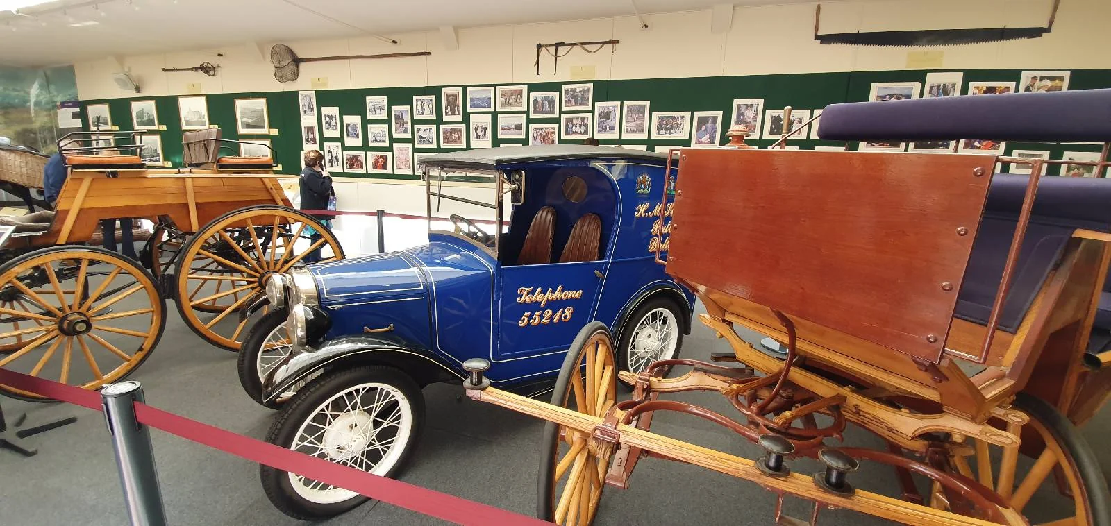 A museum exhibit featuring vintage vehicles, including a blue car labeled "Libelle" with the year 1930. The background displays framed photos on a green wall, and there are other wooden carriages with large spoked wheels.