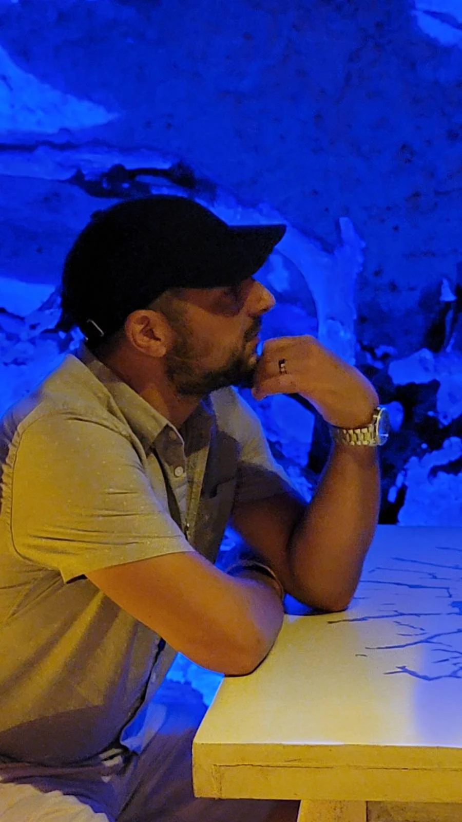 A person wearing a black cap and beige shirt sits at a table with their chin resting on their hand, looking contemplative. The background is illuminated with a blue light, creating a calm and serene ambiance.