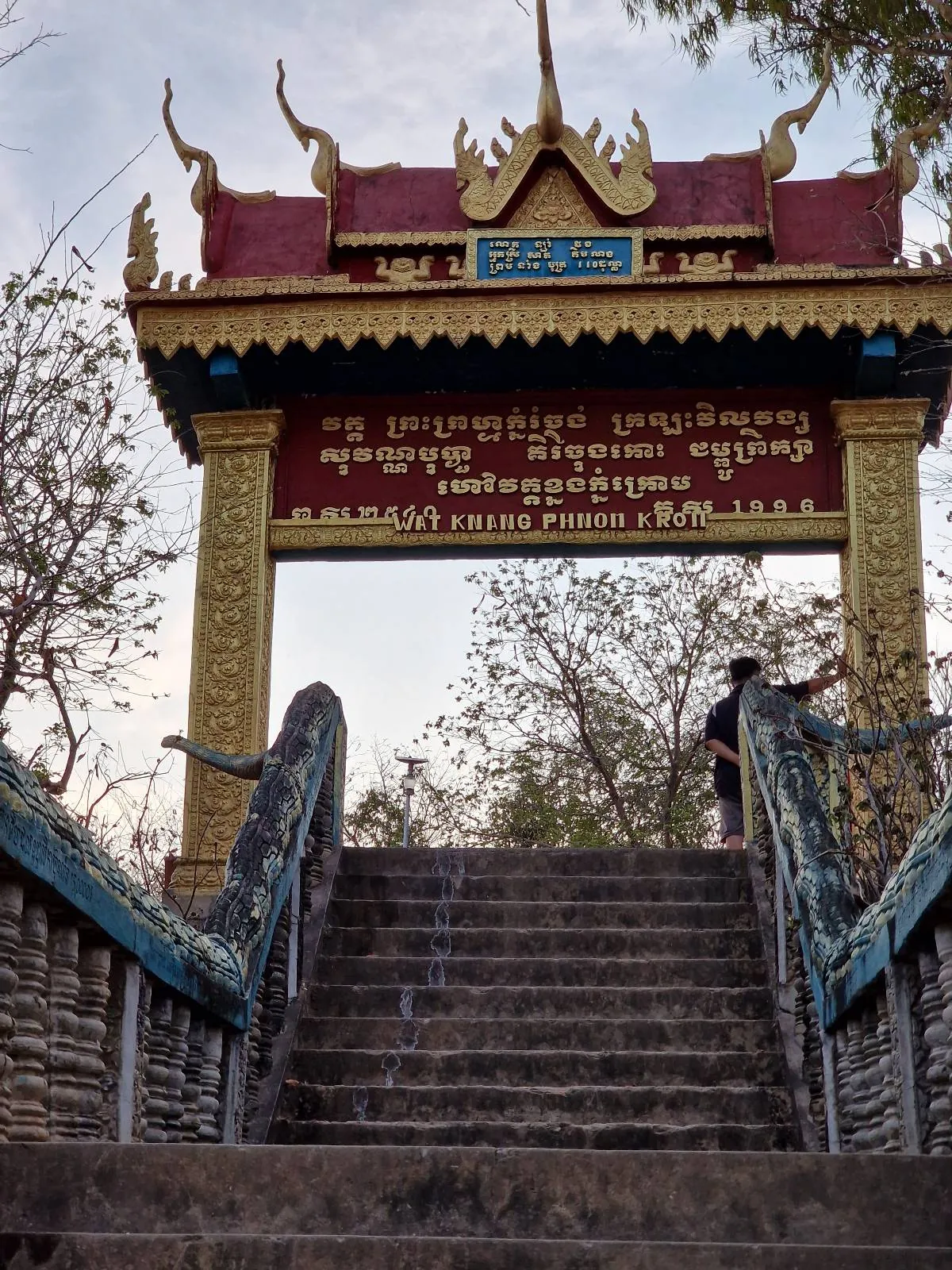 A decorative gate with golden details and script in a foreign language stands at the top of stone steps, surrounded by trees. A person is seen climbing the stairs toward the gate.