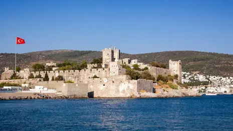 Bodrum Castle in Turkey viewed from the sea, with stone fortress walls, towers, and the Turkish flag above the coastline.