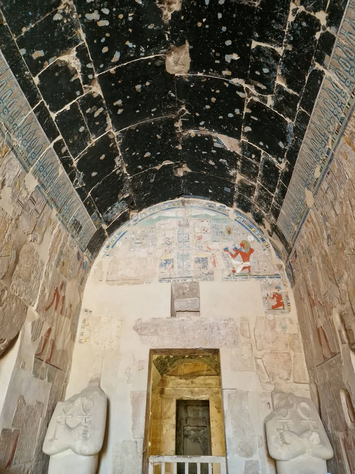 Interior of an ancient building with damaged frescoes on arched walls and a star-speckled ceiling, leading to a doorway.
