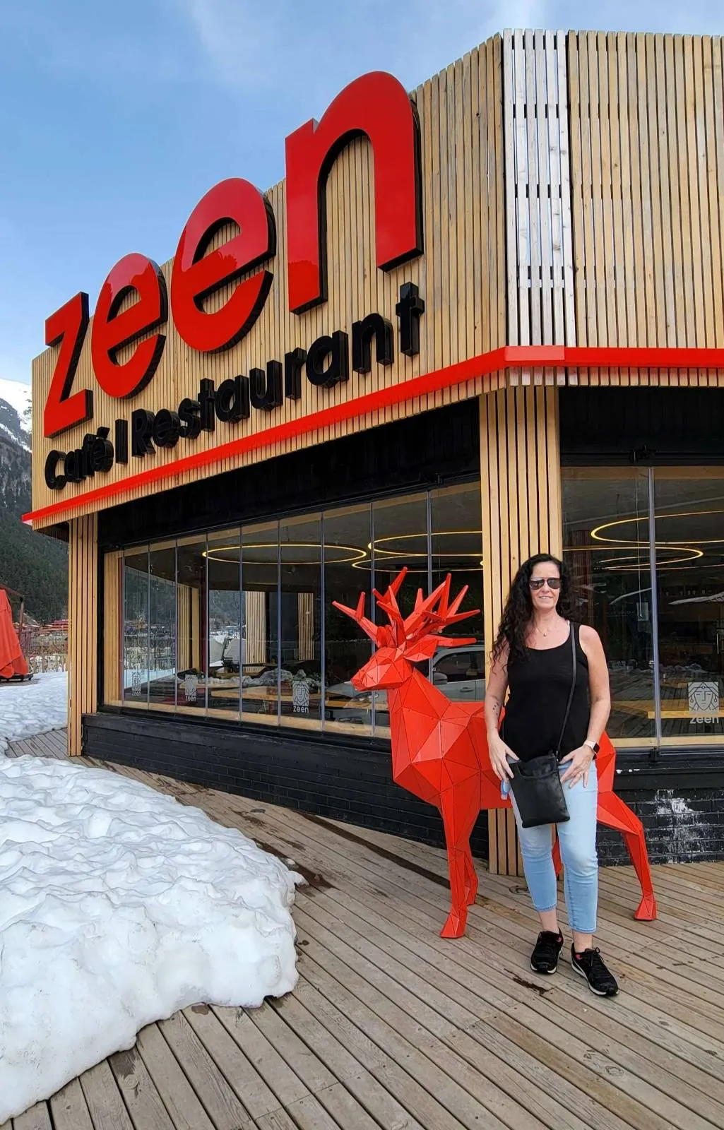 A person stands in front of Zeen Café Restaurant with a red deer statue nearby. Snow is visible on the ground.