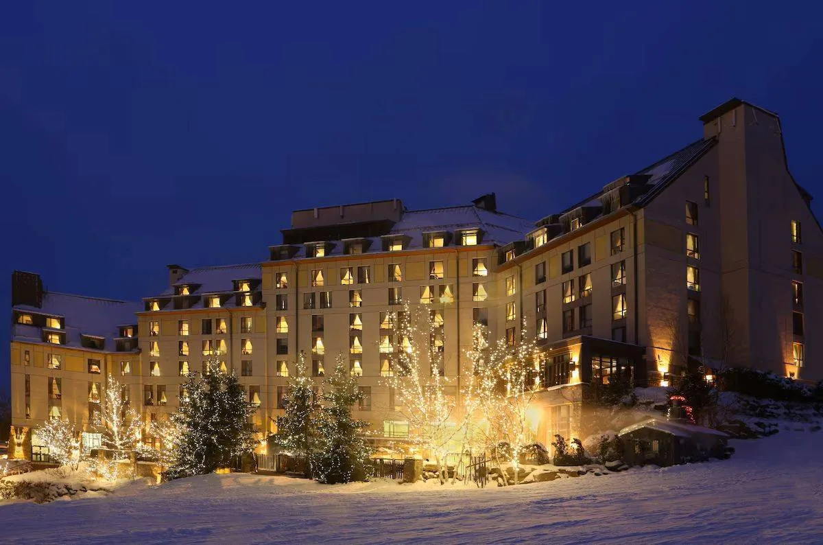 A large, illuminated hotel building in a snowy landscape under a deep blue evening sky. The warm lights from the windows contrast with the snow on the ground and roofs, creating a cozy and inviting winter scene.