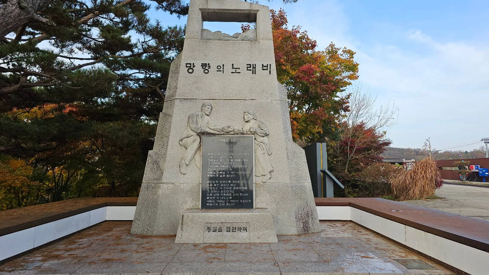 A stone monument with Korean text and engraved figures of two people facing each other. Trees with autumn foliage are in the background, and a clear sky above. A small plaque is mounted on the front.