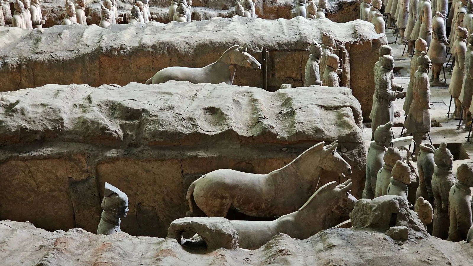 Rows of life-sized terracotta horses and soldiers stand in an ancient excavation site, partially surrounded by earth and clay walls, representing China’s famous Terracotta Army.