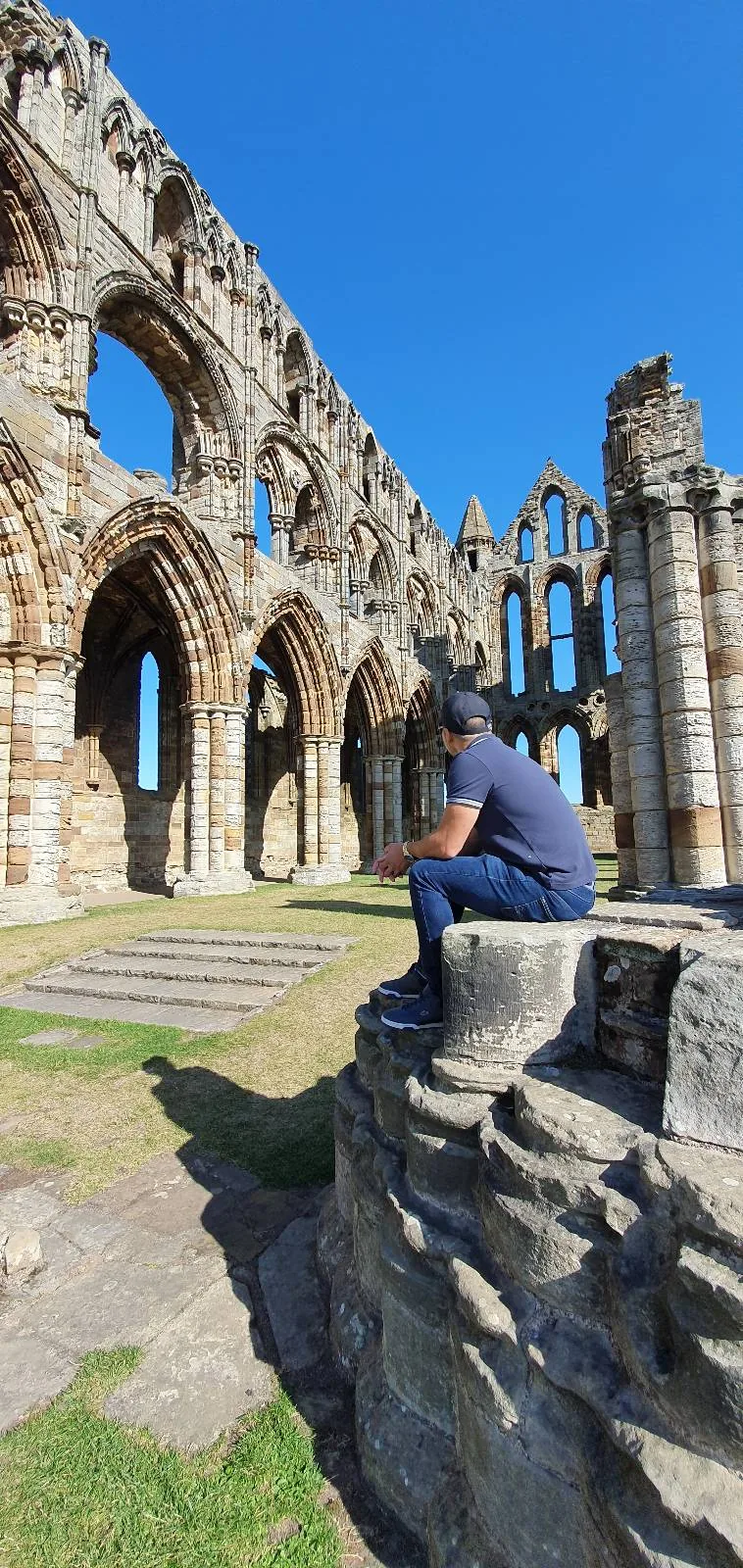 A person in a blue outfit sits on a stone ledge, with their back to the camera, gazing at the ruins of an ancient stone structure under a clear blue sky. The arches and stonework create a dramatic visual.