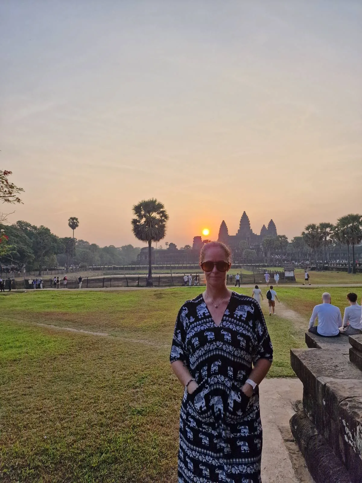 A person wearing a patterned dress stands outdoors at sunrise with the silhouette of Angkor Wat temple in the background, surrounded by green grass and a few people sitting nearby.