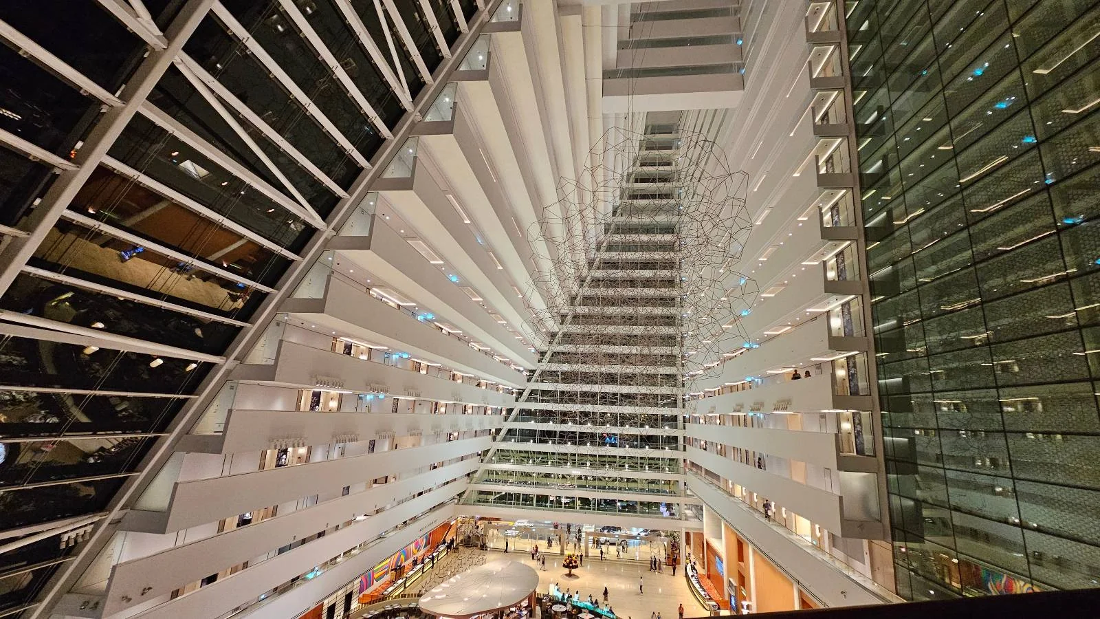 A wide-angle view of an atrium in a modern building, featuring geometric architectural lines and patterns. The perspective focuses upward, showcasing balconies and glass elements, with people gathered on the ground floor below.