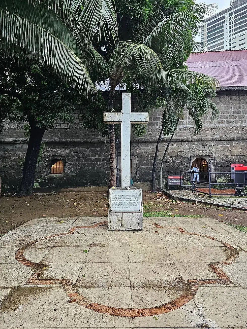 White cross on stone platform, surrounded by palm trees, next to an old stone building. Plaque with text in front. Overcast sky.