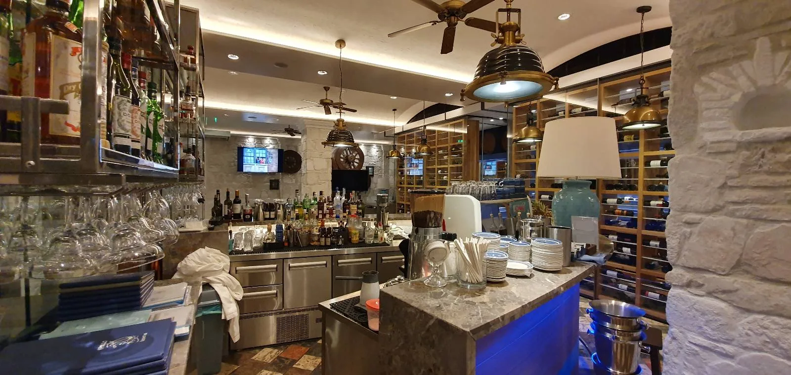 A cozy bar with a well-stocked counter and an assortment of glasses. Shelves with bottles line the walls. Warm lighting creates an inviting atmosphere. A modern lamp and industrial-style pendant lights add a stylish touch.