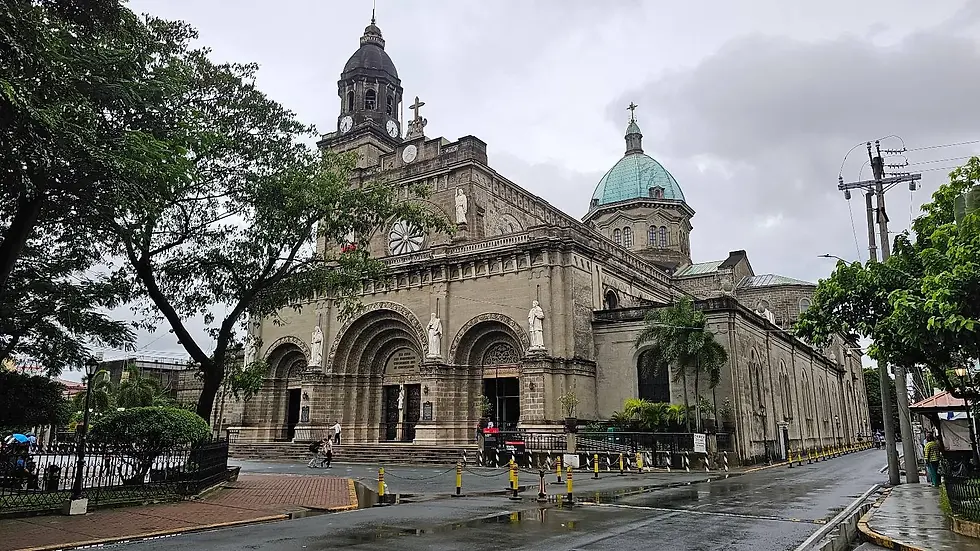 Historic cathedral with arched entrances, statues, and a central dome under a cloudy sky. Trees and a wet street in the foreground.