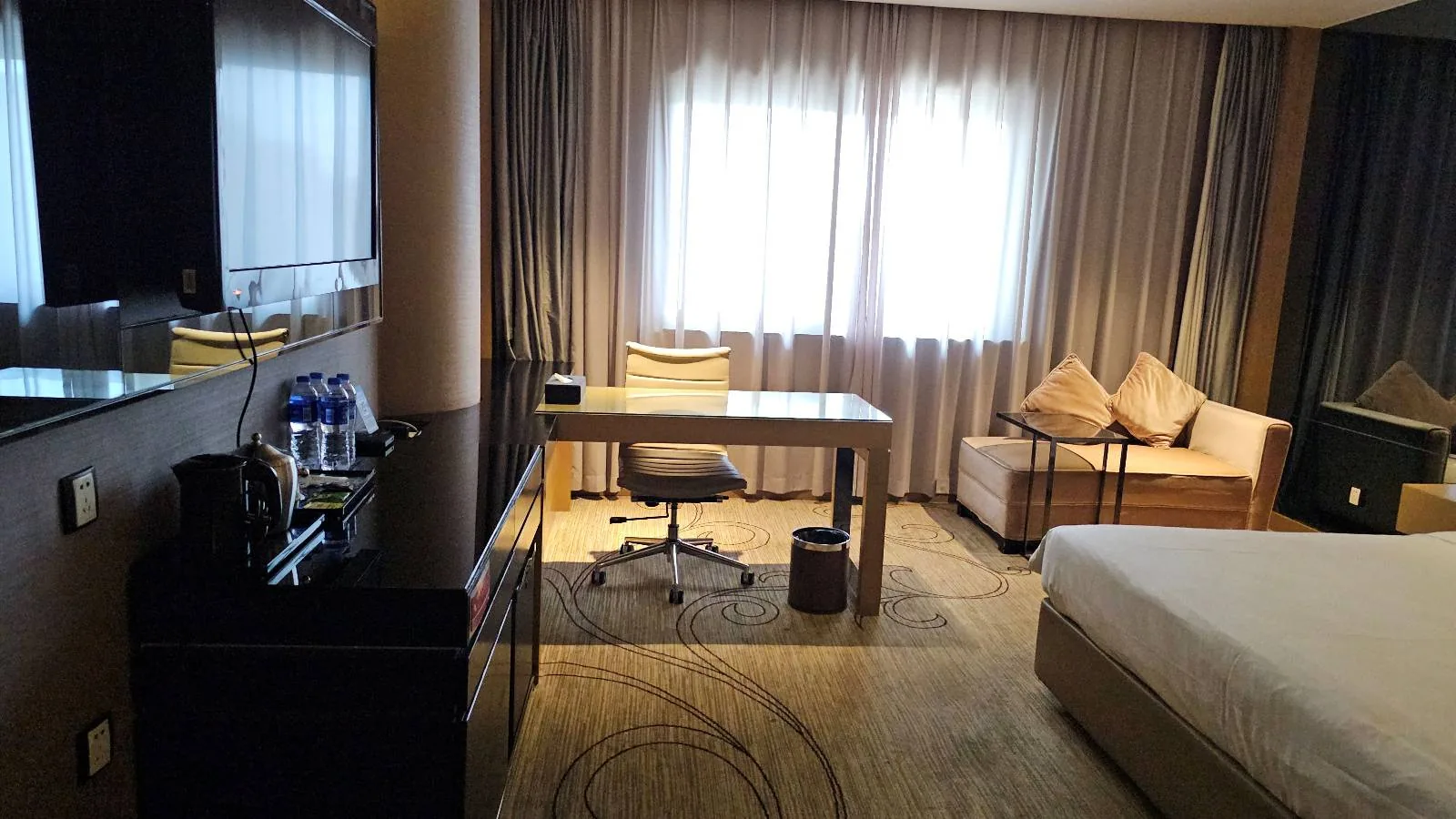 A modern hotel room features a work desk with a chair, a sofa with cushions, a bed, and a minibar with bottled water, all set against large windows with sheer curtains.