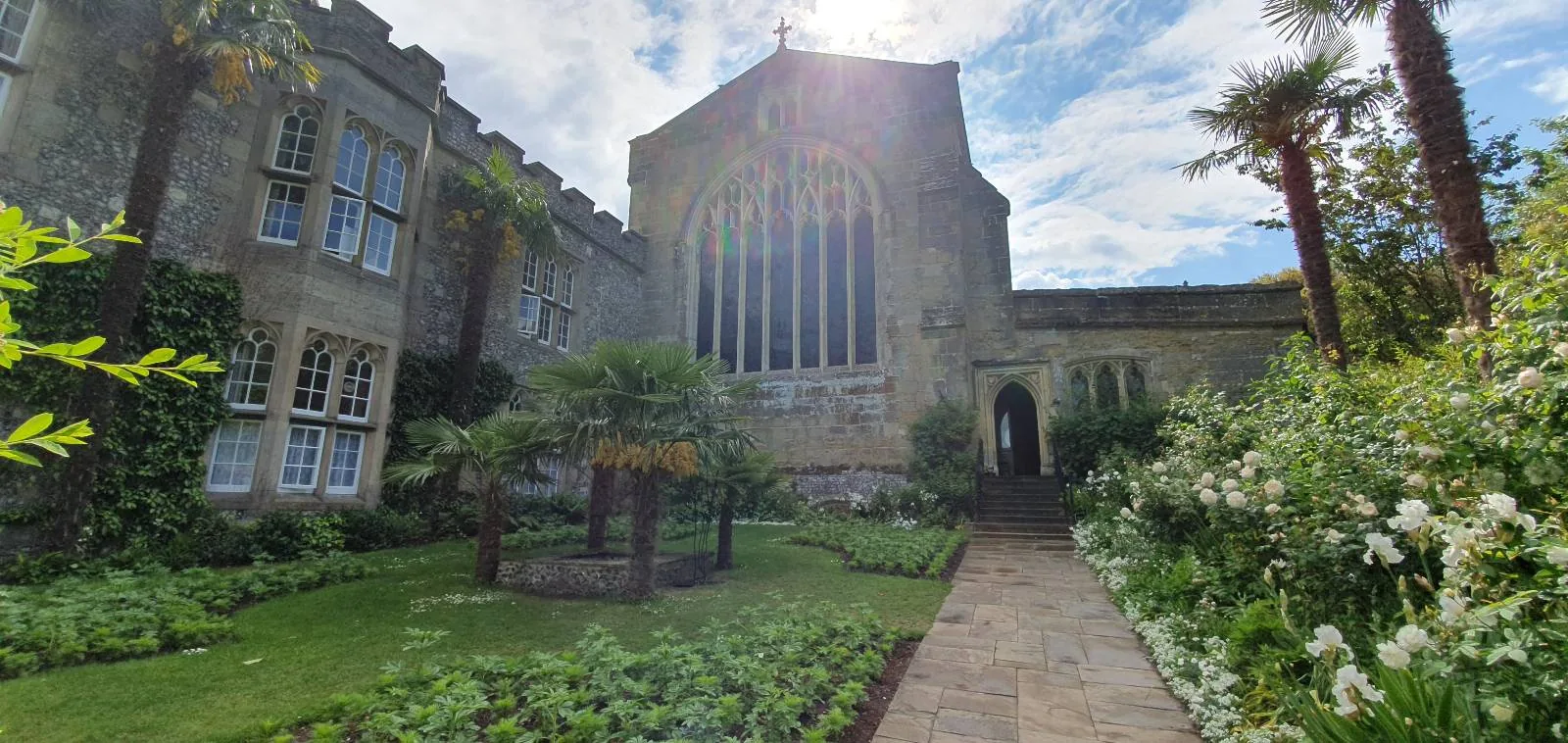 A stone building with large arched windows faces a garden with green plants, palm trees, and a stone pathway under a bright, partly cloudy sky.