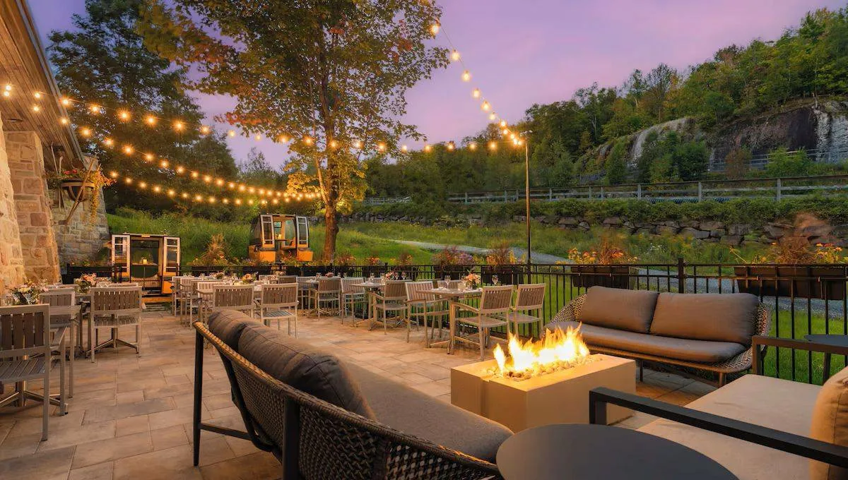 Outdoor patio at dusk with string lights and a lit fire pit. Several tables and chairs are arranged around the patio, offering a cozy atmosphere. A natural landscape with trees and rocky formations is visible in the background under a purple sky.