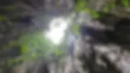 View from inside a cave looking up at a circular opening in the ceiling, surrounded by jagged rocks and green foliage against a bright sky.