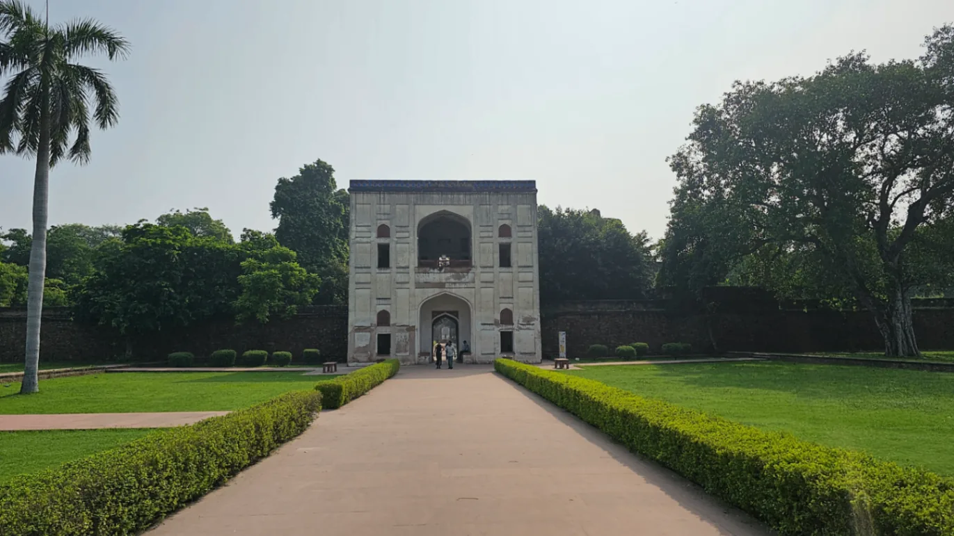 A historic stone building with arched windows stands at the end of a pathway bordered by manicured hedges. Lush green trees surround the structure under a partly cloudy sky.